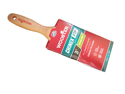 A wooden-handled brush with a red, green, and white Wooster Chinex FTP 3-inch product packaging, placed diagonally on a white surface.